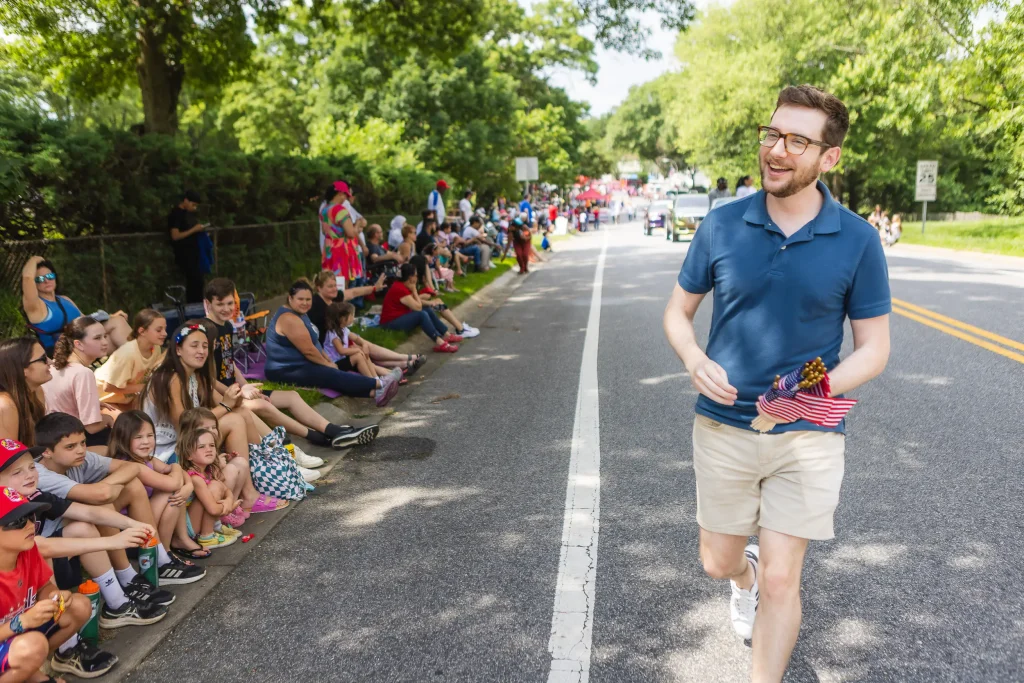 Michael Estève, mayor of Bowie Maryland, is walking down a suburban street in a parade. He is holding miniature American flags and smiling and citizens seated on the side of the road.
